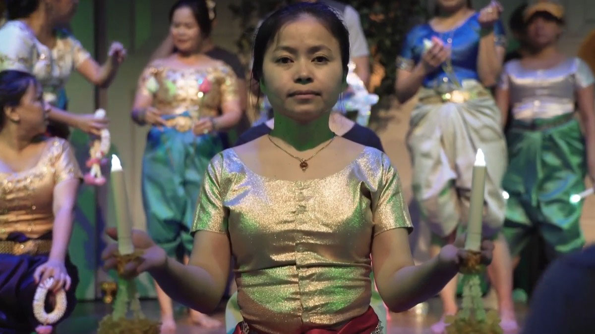 Dancers on stage in shimmering green, white, and gold costumes. A woman in front looks straight into the camera, holding a battery-powered candle in each hand.