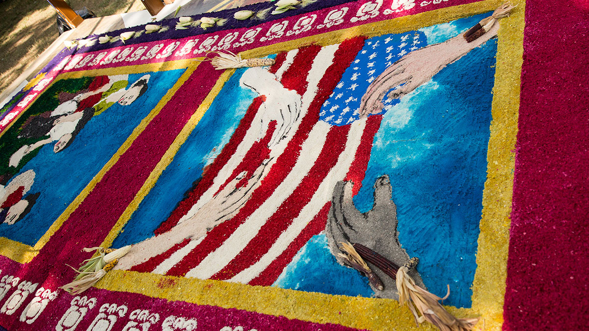 A design made of colored sawdust laid out on the ground, showing four hands of different skin tones around an American flag.