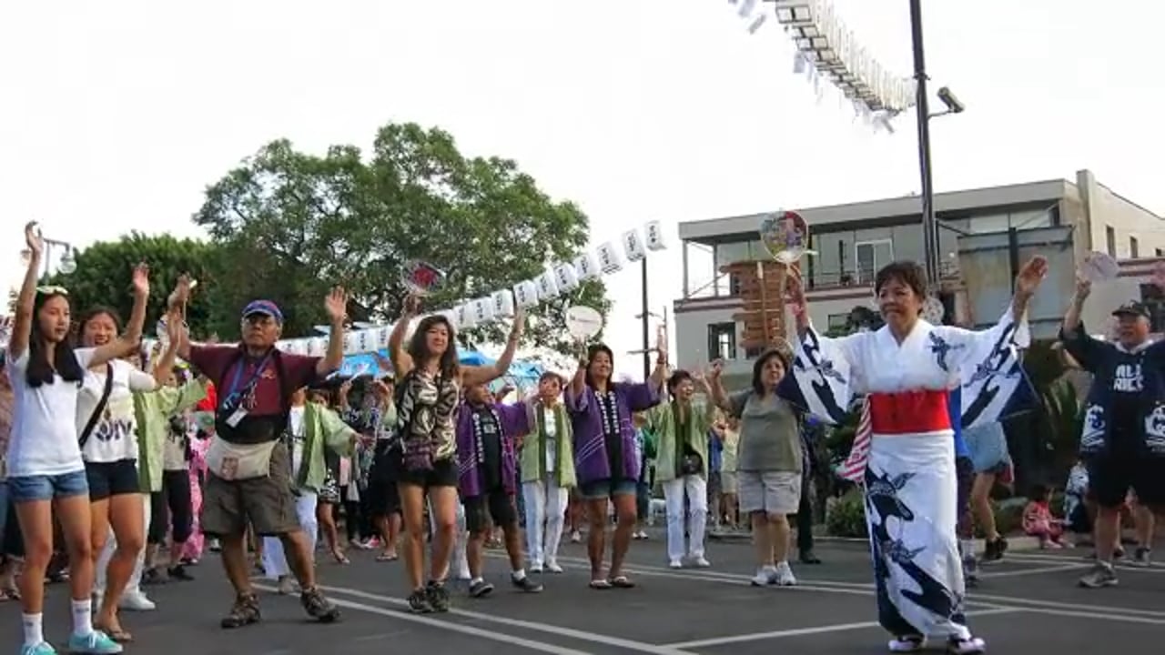 Dancers in a parking lot, arms raised, some holding paper fans.